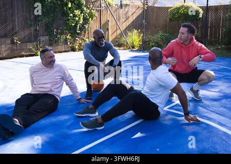 Diverse male friends sitting and talking on blue backyard court with basketball in hoodies sneakers Stock Photo
