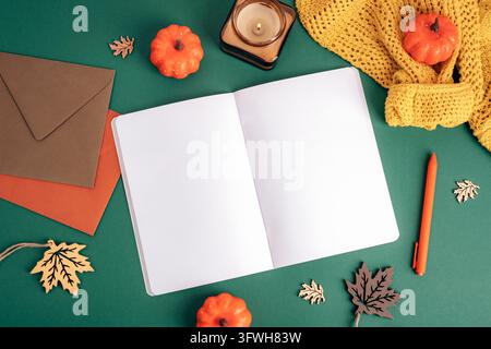 Open blank notebook with an orange pen, envelopes, pumpkin decorations, candle and yellow knitted scarf on a green background. Autumn seasonal Stock Photo