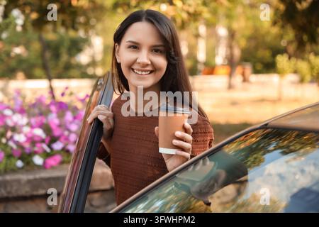 Commuter lifestyle and morning routine, happy woman holding a coffee cup while getting out of her car in a park setting for a daily commute journey Stock Photo