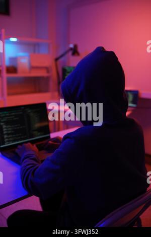 A mysterious hooded hacker working on a computer in a dark room. A concept for cybersecurity, hacking, cybercrime, and data privacy threats. Stock Photo