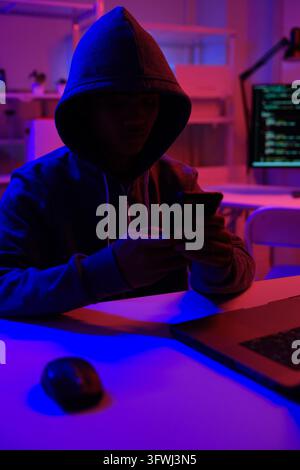 A mysterious hooded hacker working on a computer in a dark room. A concept for cybersecurity, hacking, cybercrime, and data privacy threats. Stock Photo