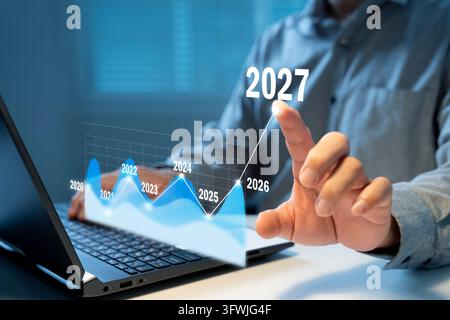 Hand taps glowing 2027 on futuristic graph. Person in gray shirt interacts with holographic data. Blue tones show growth trend from 2021 to 2027. Idea Stock Photo