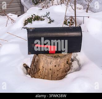 Black rural mailbox with a red flag mounted on a weathered tree stump, surrounded by deep fresh snow in a quiet winter yard. Stock Photo