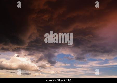 Dramatic storm clouds sweep across the sky at sunset, casting red and dark atmospheric light over the horizon and creating a moody. Stock Photo