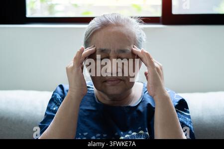 Elderly Asian woman sitting and having a headache and touching her head with her hands at home. Asian older lonely woman. Retirement, quarantine and h Stock Photo