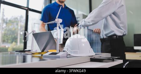 Two Asian engineers wearing hardhats and safety vests discussing solar panel installation and renewable energy project in the office. Stock Photo