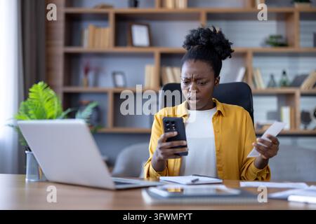 Young professional woman in office chair studies smartphone with worried expression while holding paper, seated at desk with laptop, notebook, and shelves behind her in blurred background. Stock Photo