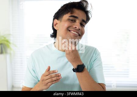 Smiling Indian man in light blue T-shirt touching chin, checking smartwatch by blinds with plant Stock Photo