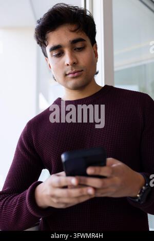 African American man in 20s holding phone, watch visible, wearing burgundy knit near glass wall Stock Photo