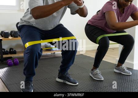 Woman and man performing resistance-band squats on black mat in small home gym with dumbbells Stock Photo