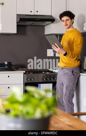 African American man wearing mustard sweater holding tablet and checking recipe by stove Stock Photo
