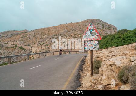 A cyclist rides a road bike around a curve on a mountain road in Pollença, Spain. A prominent triangular road sign covered in stickers stands by the r Stock Photo