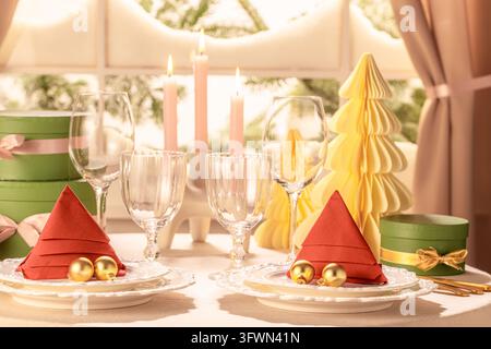 Horizontal dining table setup for two featuring red tree napkins, golden ornaments, crystal glasses, yellow paper fir trees, glowing pink candles, and Stock Photo