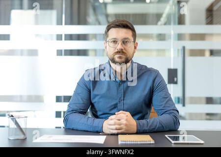 Portrait of bearded businessman seated at office desk with hands clasped, wearing glasses and blue shirt. Documents, notebook, pen, and tablet suggest planning, review, and focused professional work. Stock Photo