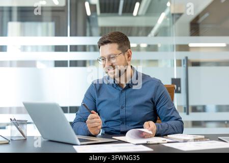 Adult man in blue shirt works at office desk with laptop, papers, notebook and pen, reviewing documents and writing notes beside glass wall in bright modern workspace during focused daily tasks. Stock Photo