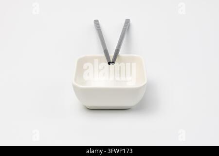 Two black textured chopsticks with tips resting inside empty small white ceramic square bowl for soy sauce. Pair forms upright V shape on plain white Stock Photo