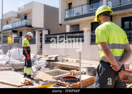 Two construction workers in high-visibility uniforms and safety helmets inspect an urban street maintenance site beside modern residential buildings. Stock Photo