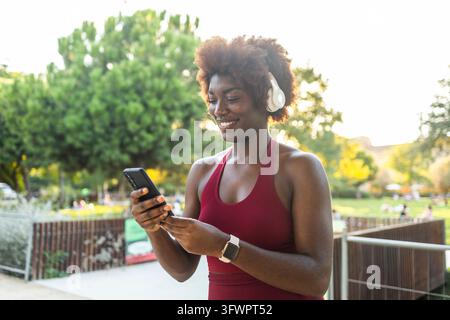 Smiling young Black woman in sportswear uses a smartphone while wearing headphones in a sunny park, taking a break from her outdoor running workout. Stock Photo