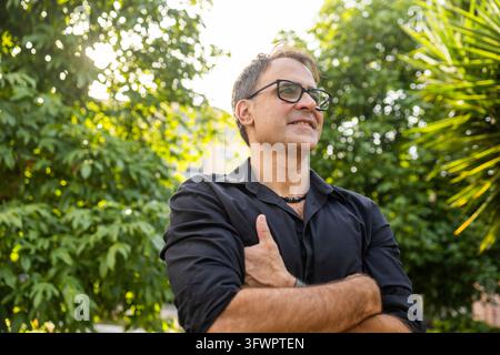 Mature man wearing glasses and a black shirt stands with crossed arms in a lush garden, smiling confidently amid sunlit tropical foliage and natural g Stock Photo