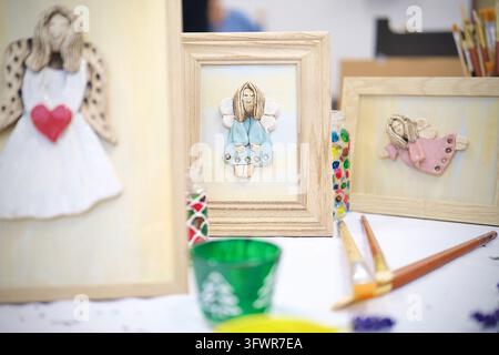 Framed angel decorations in blue, white and pink dresses displayed with paintbrushes and colourful decorated glass holders. Stock Photo