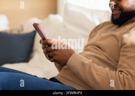 African American man reclining on light-colored sofa at home, holding pink smartphone and typing. Couch, denim, beige, casual, relaxed, cozy, text Stock Photo