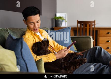 Asian man wearing yellow shirt gray pants sitting on green sofa cradling dog holding phone Stock Photo