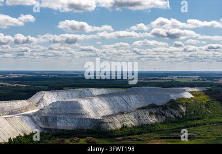Aerial view of the large open-pit mine with white soil and rock formations, surrounded by green forests and distant wind turbines under a blue sky in Stock Photo
