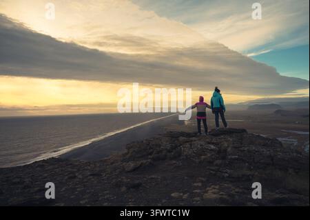 View of two people standing on the rocky cliffs of Dyrholaey overlooking the black sand beach and the Atlantic Ocean at sunset in Myrdalshreppur, Icel Stock Photo