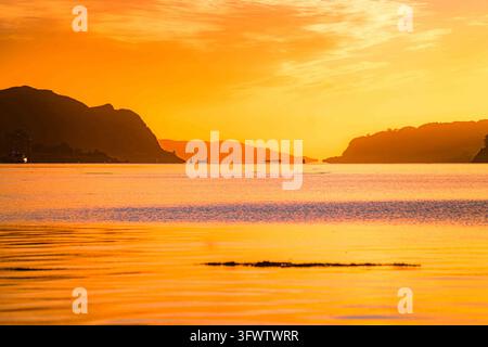View of the sunset over the bay with silhouettes of small boats and distant mountains under a golden sky in Oban, Scotland, United Kingdom. Stock Photo