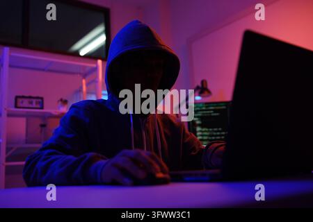 A mysterious hooded hacker working on a computer in a dark room. A concept for cybersecurity, hacking, cybercrime, and data privacy threats. Stock Photo