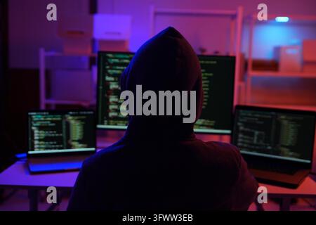 A mysterious hooded hacker working on a computer in a dark room. A concept for cybersecurity, hacking, cybercrime, and data privacy threats. Stock Photo