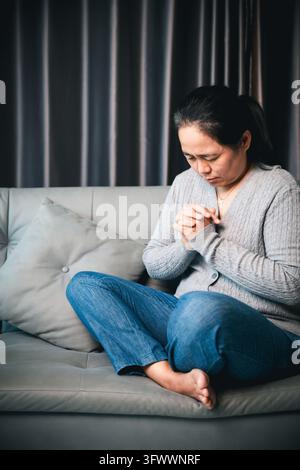A woman wearing a gray cardigan and cross necklace sits with legs folded on a sofa, head bowed and hands clasped near her chest, leaving space on the Stock Photo