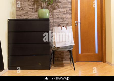Canvas tote bag resting on a chair beside a dark cabinet and wooden door in a home entryway Stock Photo