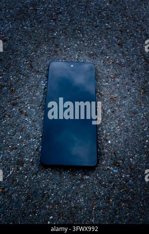 Black smartphone lying face up on rough asphalt, its intact dark screen reflecting a cloudy sky in a top-down outdoor photograph Stock Photo