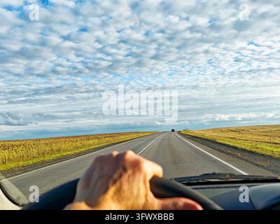 Driver viewpoint of hand on steering wheel driving down long straight rural road between grassy fields under expansive cloudy sky, low sun warm tones, Stock Photo