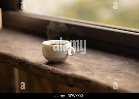 Ha Giang, Vietnam - January 13, 2026: A white ceramic cup filled with steaming black coffee resting on a rustic wooden windowsill. Stock Photo