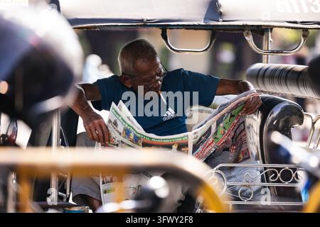 Chiang Mai, Thailand - January 17, 2026: An older man wearing glasses and a blue shirt reads a newspaper while seated inside a tuk-tuk vehicle. Stock Photo