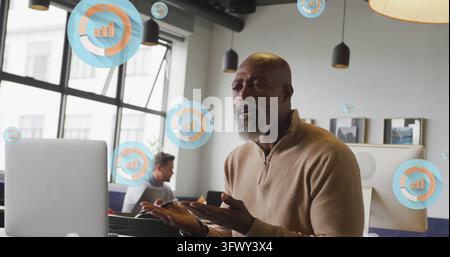 Image of multiple bar graph icons over african american man having imagecall on laptop at office. Computer interface and business data technology conc Stock Photo