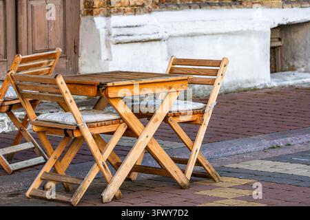 A set of wooden folding chairs with fabric cushions surrounds a matching wooden table on a paved sidewalk outside an old building. The whitewashed bas Stock Photo