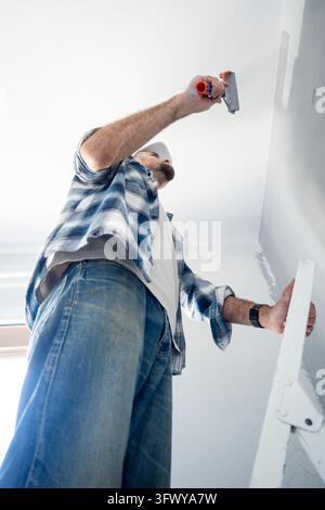 Low angle view of a man standing on a ladder and using a small paint roller to reach the upper section of an interior wall during home renovation. Stock Photo