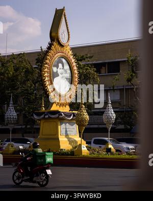 Bangkok, Thailand - February 2, 2026: A delivery rider passes a large golden memorial portrait on a city street. Stock Photo