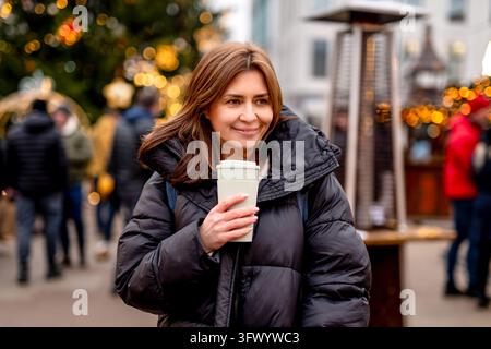Smiling woman holding hot coffee in festive Christmas market lights during winter evening Stock Photo