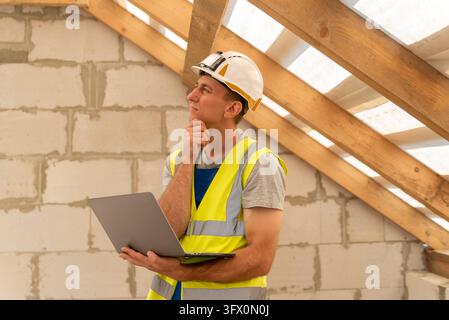 Construction worker wearing a hard hat and safety vest, holding a laptop and contemplating plans in an unfinished building with wooden roof beams and Stock Photo