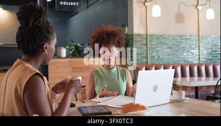Discussing women in business-casual clothing gesturing working at cafe table laptop tablet coffee Stock Photo