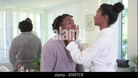African American couple wearing bathrobes, man sitting on vanity, woman placing hands on man's face. Skincare, grooming, partners, bathroom, mirror, s Stock Photo