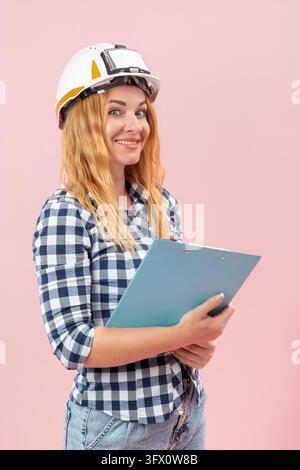 Young woman smiling while wearing a white safety hardhat and blue plaid shirt, confidently holding a blank clipboard, representing expertise and proje Stock Photo