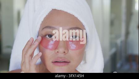 Asian woman wearing towel applying under eye beauty mask in sunny bathroom Stock Photo