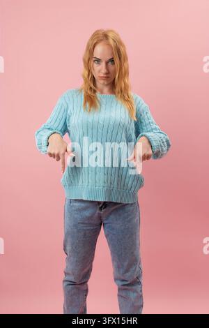 Young woman in a light blue sweater and jeans stands against a pink background, pointing her fingers downwards with a stern, concentrated facial expre Stock Photo