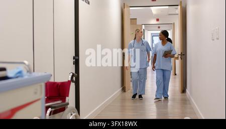 Diverse female nurses walking and talking down hospital corridor in blue scrubs, stethoscope tablet Stock Photo