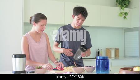 Diverse couple preparing fruit at kitchen island, woman slicing strawberries, man pouring blender Stock Photo
